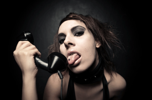 An image picturing a young woman with a punk-like aesthetic, with a pierced tongue, about to lick the receiver of a black old fashioned telephone
